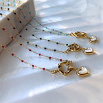 Four beaded necklaces in different colors, each with a gold toggle clasp and heart-shaped mother of pearl charm, arranged diagonally on a marble-effect surface with soft natural lighting.