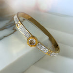 Gold bangle bracelet with central zirconia stone, surrounded by rows of shimmering crystals, placed on a soft white jewelry stand.