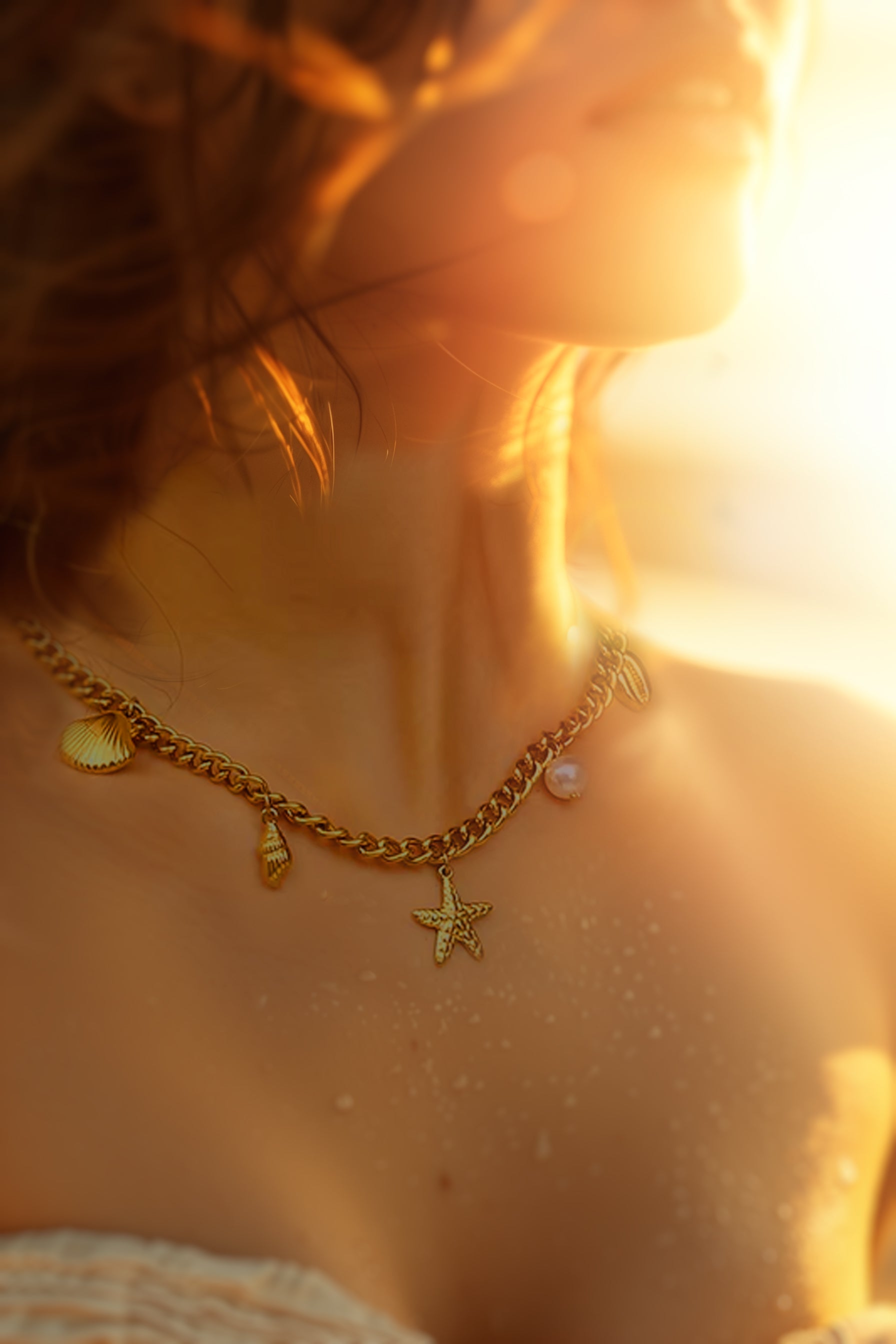 A woman bathed in golden sunset light, wearing a gold necklace with shell and starfish charms — symbolic jewelry from the Water collection, inspired by the calm and power of the ocean.