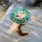 A beaded necklace with translucent turquoise-blue glass beads and a textured gold crescent moon pendant, displayed on a white satin fabric under warm, dappled sunlight.