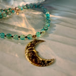 A beaded necklace with translucent turquoise-blue glass beads and a gold crescent moon pendant engraved with floral and eye details, resting on a white textured surface with warm sunlight shadows.