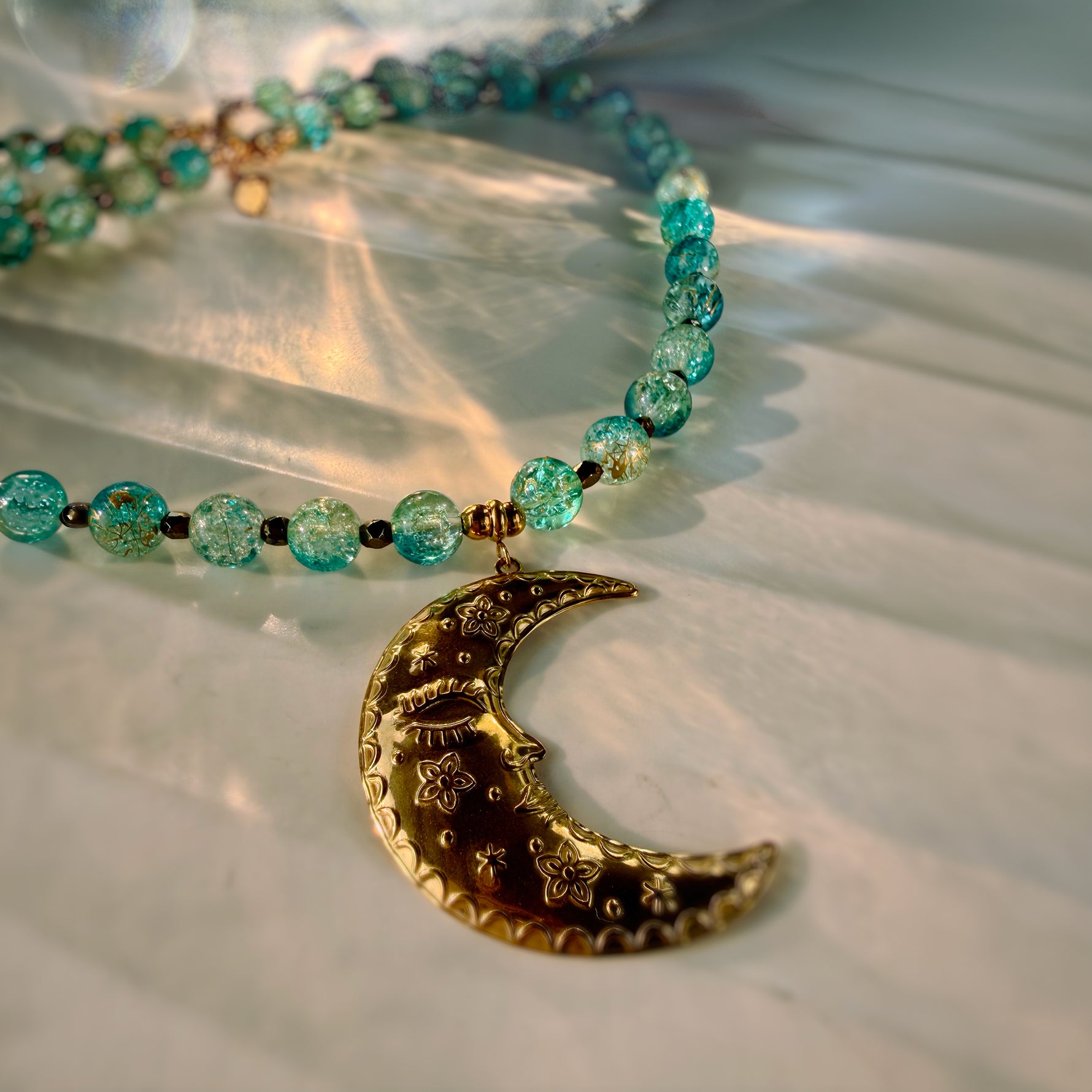 A beaded necklace with translucent turquoise-blue glass beads and a gold crescent moon pendant engraved with floral and eye details, resting on a white textured surface with warm sunlight shadows.