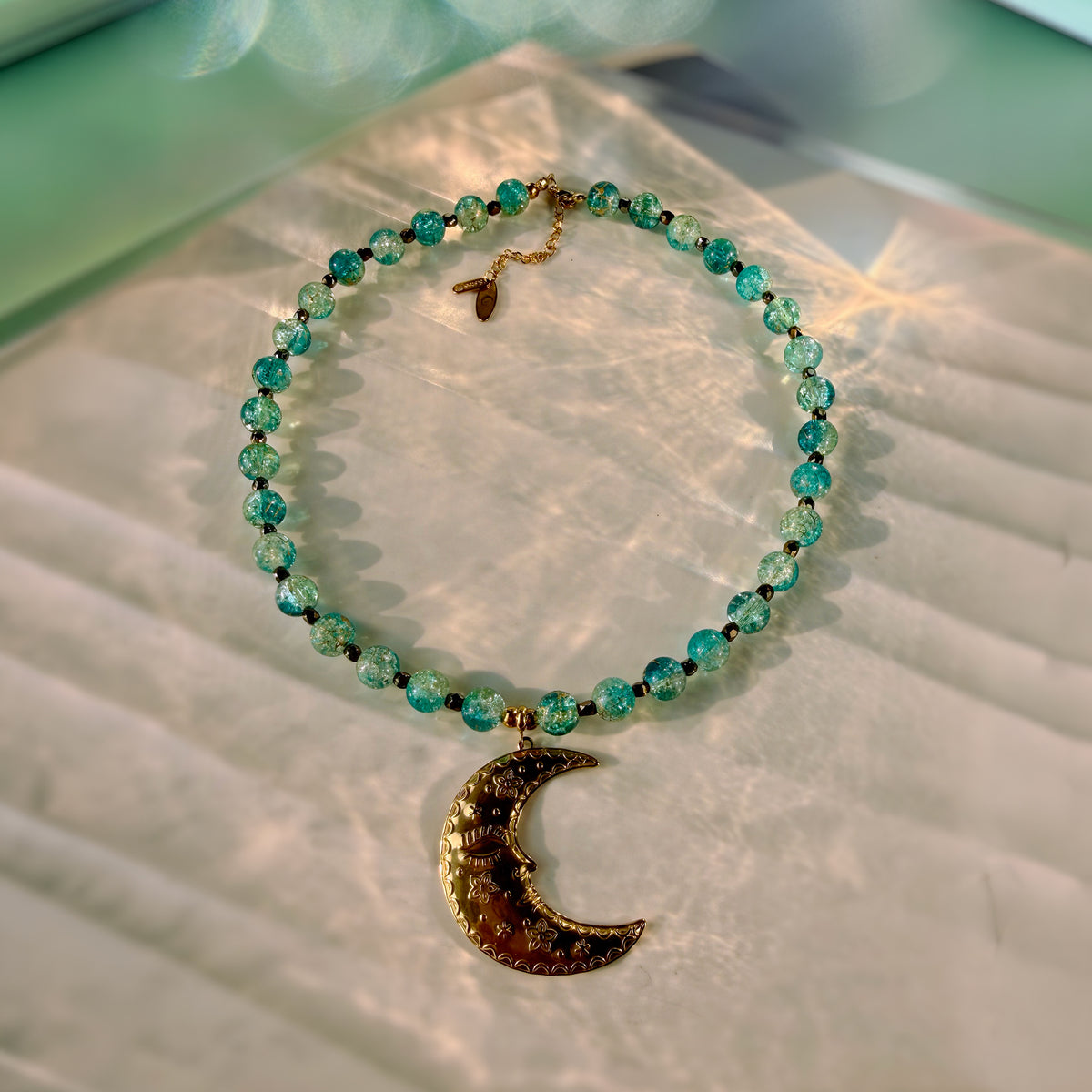 A beaded necklace with faceted turquoise and green beads, featuring a gold crescent moon pendant embellished with small rhinestones, displayed on a textured white surface.