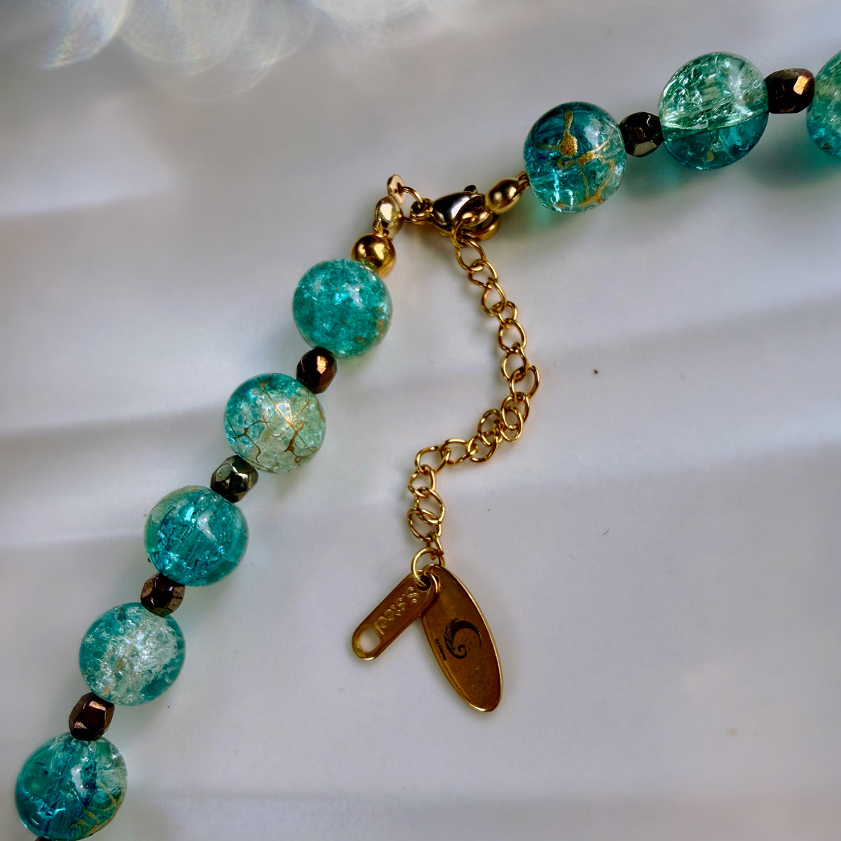 A close-up of a beaded necklace with round turquoise-blue marbled beads, small dark metallic spacer beads, and gold-toned chain hardware including a lobster clasp, extender chain, and two tags.