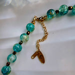 A close-up of a beaded necklace with round turquoise-blue marbled beads, small dark metallic spacer beads, and gold-toned chain hardware including a lobster clasp, extender chain, and two tags.