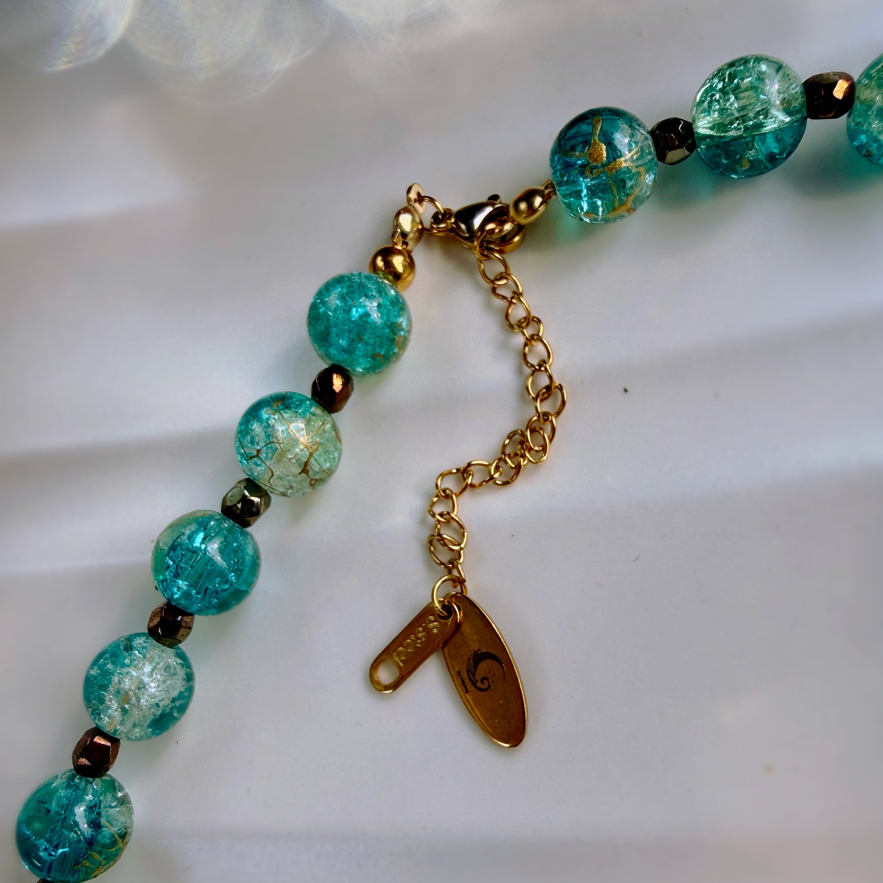 A close-up of a beaded necklace with round turquoise-blue marbled beads, small dark metallic spacer beads, and gold-toned chain hardware including a lobster clasp, extender chain, and two tags.