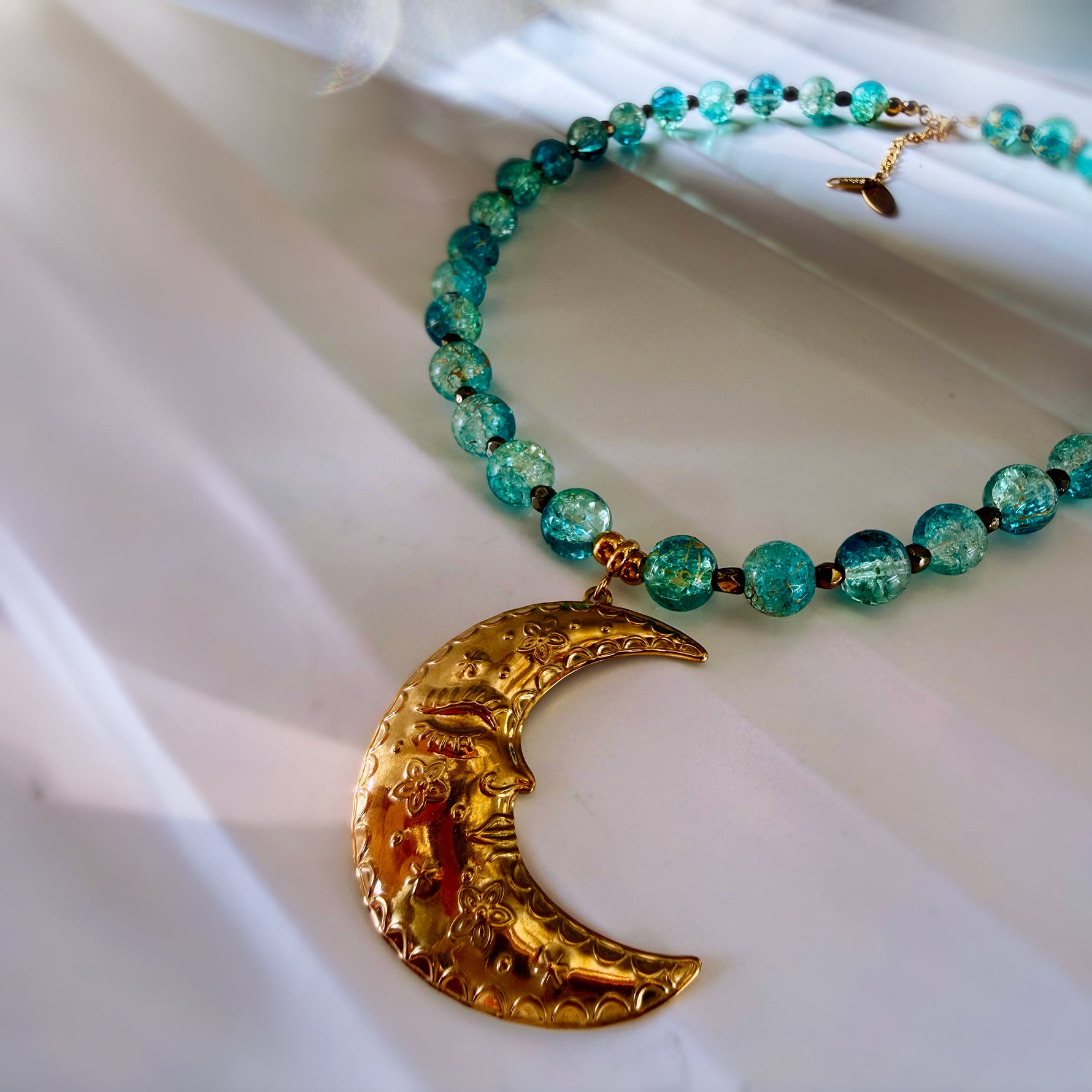 A beaded necklace with translucent teal and blue-green beads, accented with small gold spacers and a large textured gold crescent moon pendant, displayed on a white draped fabric background.