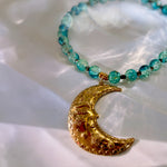 A beaded necklace with translucent turquoise-blue beads and a gold crescent moon pendant featuring a sleeping face and floral details, displayed on a white draped fabric background.
