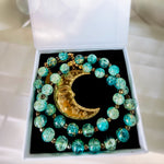 A beaded necklace with turquoise and green marbled beads and a gold crescent moon pendant, displayed inside an open white jewelry box with a black insert.
