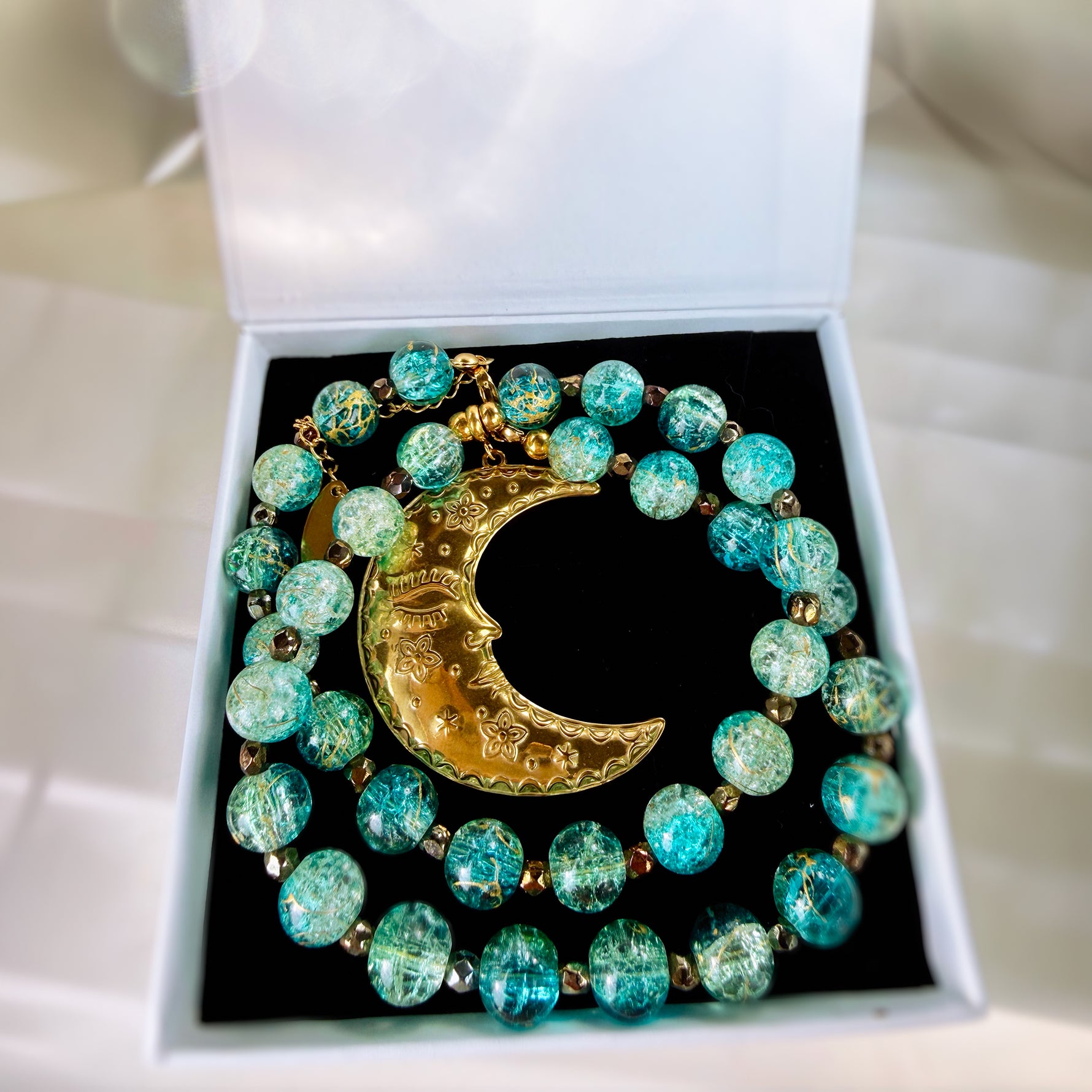 A beaded necklace with turquoise and green marbled beads and a gold crescent moon pendant, displayed inside an open white jewelry box with a black insert.