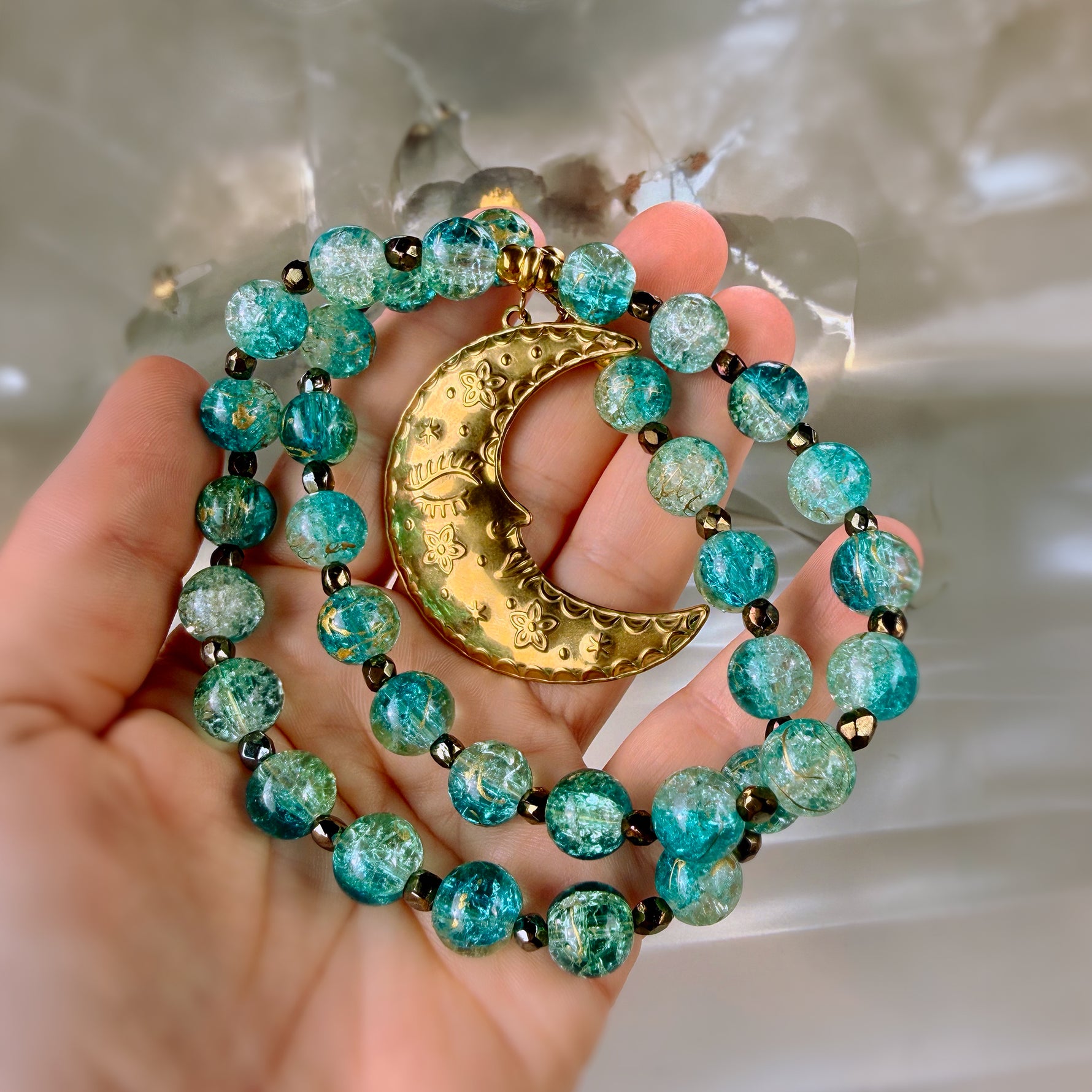 A close-up of a hand holding a beaded necklace with turquoise-blue marbled beads, small dark spacer beads, and a gold crescent moon pendant with a sleeping figure design.