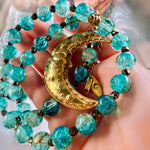 A close-up of a hand holding a beaded necklace with translucent turquoise-blue beads, small black spacer beads, and a large gold crescent moon pendant with floral engravings.