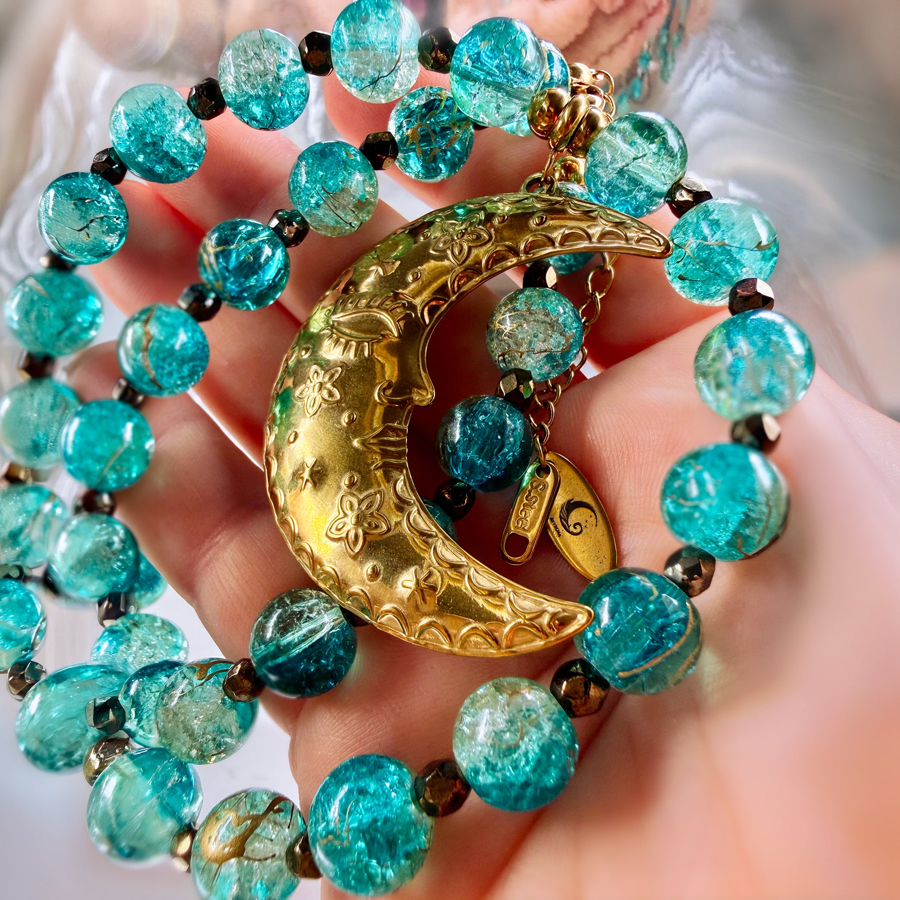 A close-up of a hand holding a beaded necklace with translucent turquoise-blue beads, small black spacer beads, and a large gold crescent moon pendant with floral engravings.
