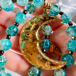 A close-up of a beaded necklace with a crescent moon pendant held in a hand. The necklace features textured turquoise-blue glass beads and a polished gold crescent moon charm with floral engravings and a small dangling tag.