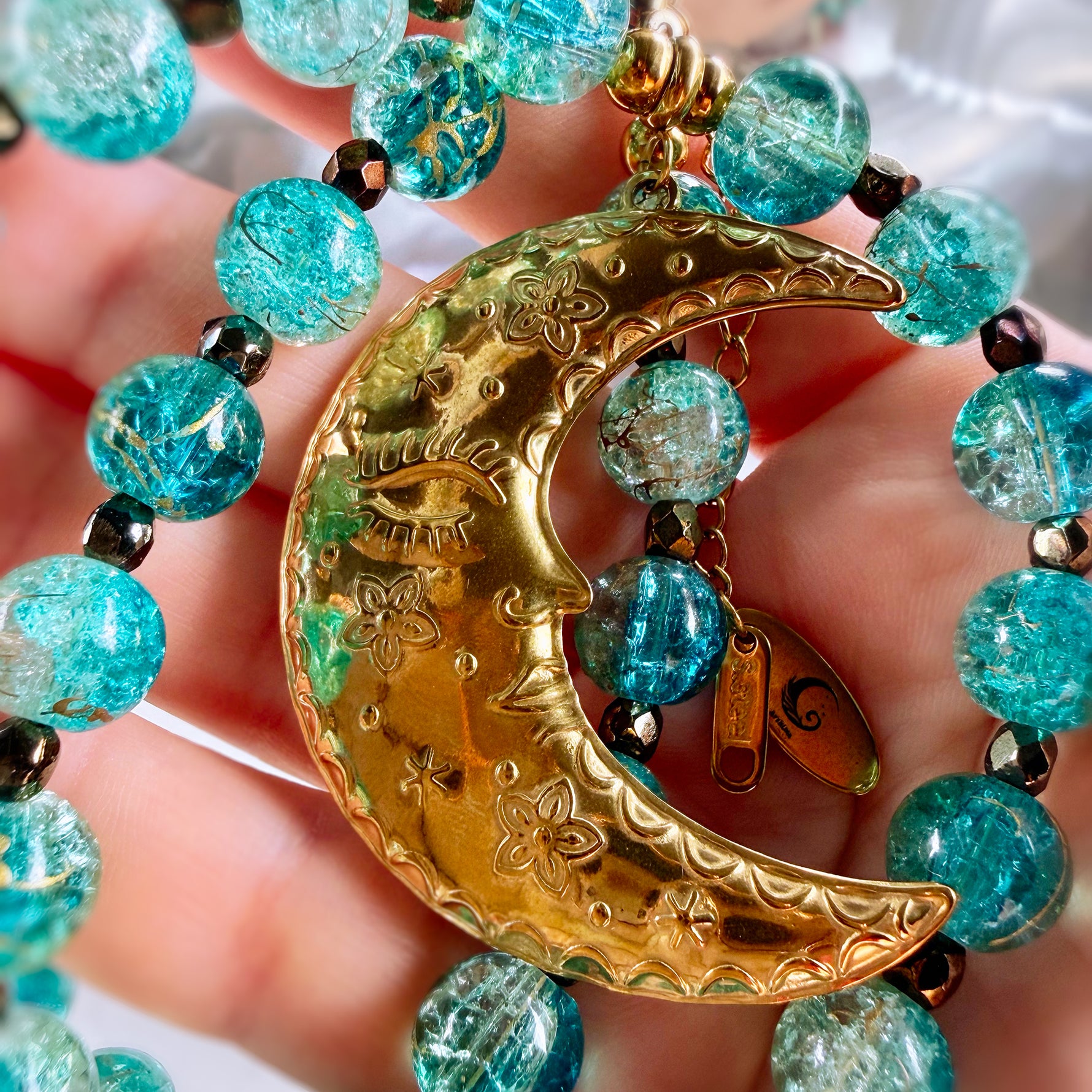 A close-up of a beaded necklace with a crescent moon pendant held in a hand. The necklace features textured turquoise-blue glass beads and a polished gold crescent moon charm with floral engravings and a small dangling tag.