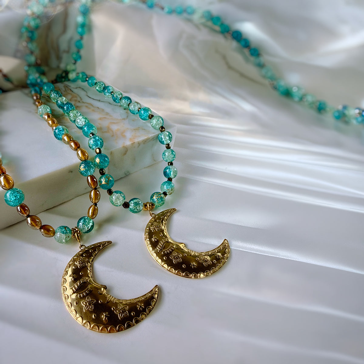 A close-up of a bohemian-style beaded necklace with turquoise and green beads, brown metallic beads, and two textured gold crescent moon pendants, styled on a white satin fabric and a marble block.