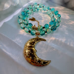 A beaded necklace with translucent turquoise-blue glass beads and a large gold crescent moon pendant, photographed on a soft white fabric surface with warm sunlight and shadows.