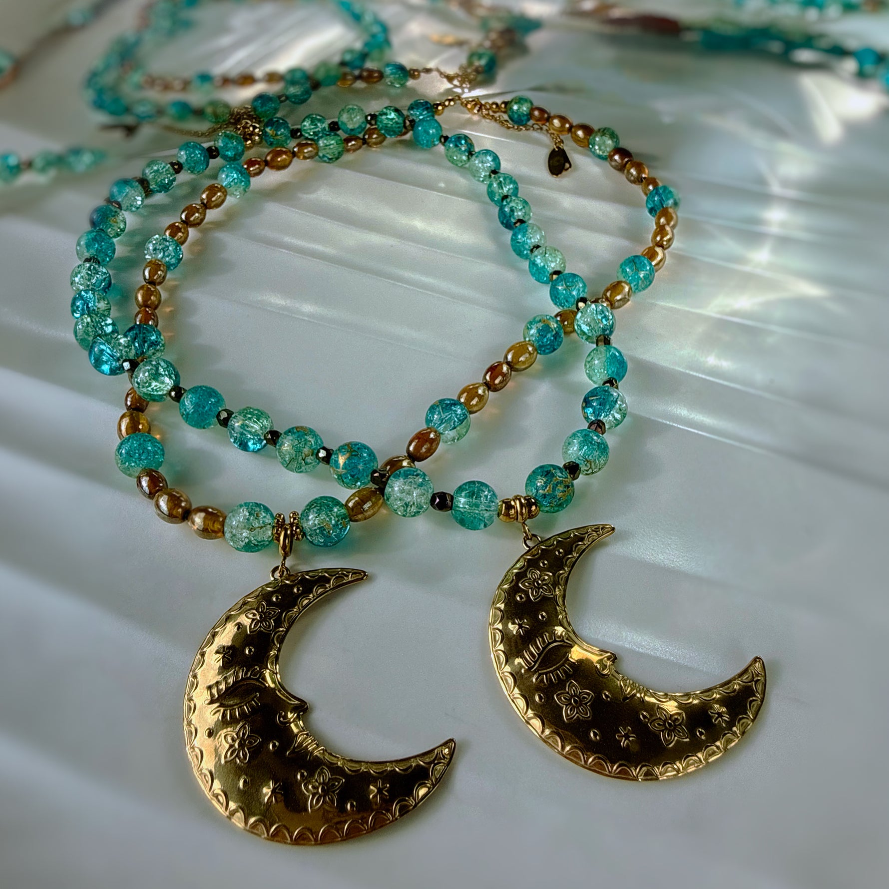 A close-up of a beaded necklace with turquoise-blue faceted beads, small metallic gold spacer beads, and two large textured gold crescent moon pendants, arranged on a light reflective surface.