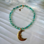 A beaded necklace with turquoise-blue marbled beads and a textured gold crescent moon pendant, displayed on a white textured fabric background.