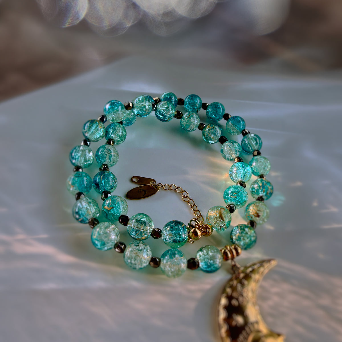 A beaded necklace with translucent turquoise-blue glass beads, small dark spacer beads, a gold-toned chain extender, and a dangling gold-toned crescent moon charm, photographed on a soft white fabric surface with warm light reflections.