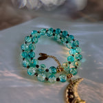 A beaded necklace with translucent turquoise-blue glass beads, small dark spacer beads, a gold-toned chain extender, and a dangling gold-toned crescent moon charm, photographed on a soft white fabric surface with warm light reflections.