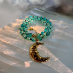 A beaded necklace with translucent turquoise-blue beads and a textured gold crescent moon pendant, styled on a translucent white surface with warm sunlight and bokeh.