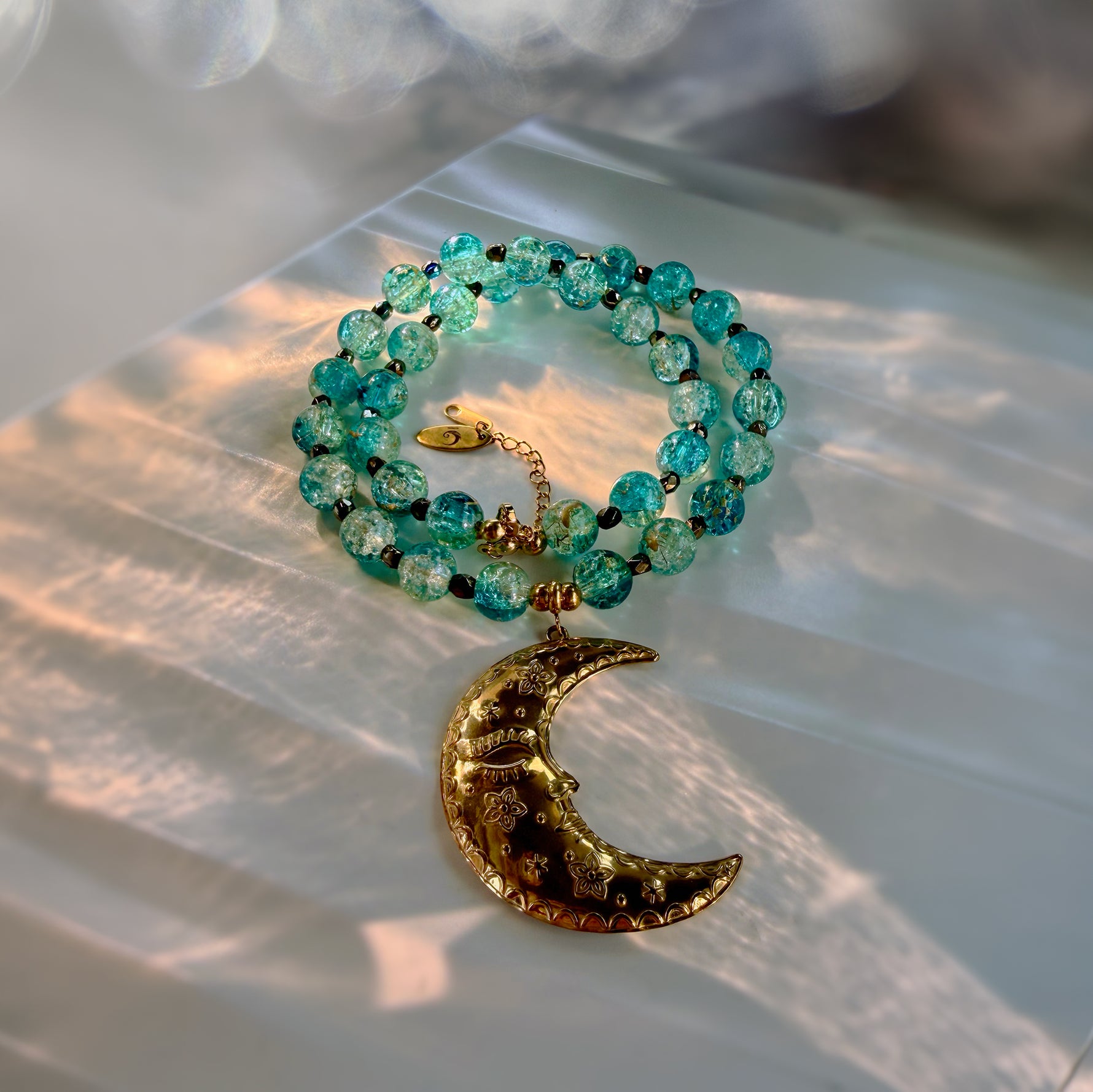 A beaded necklace with translucent turquoise-blue beads and a textured gold crescent moon pendant, styled on a translucent white surface with warm sunlight and bokeh.