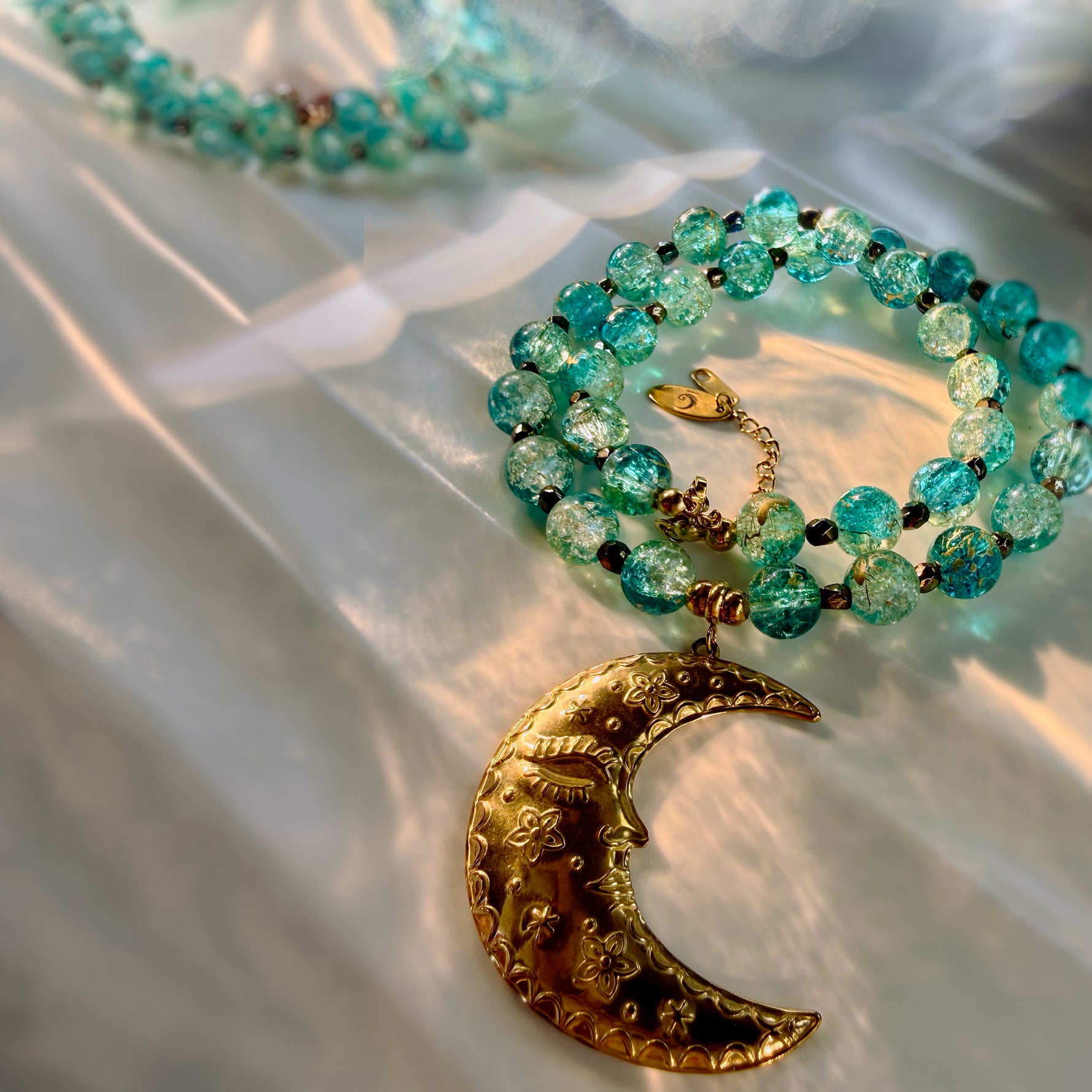 A beaded necklace with translucent turquoise-blue beads and a large gold crescent moon pendant, arranged on draped white fabric.