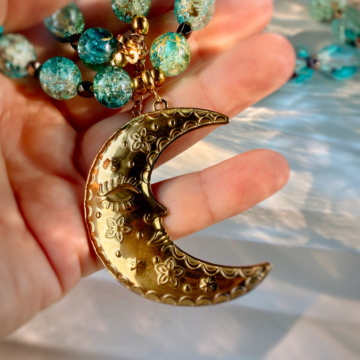 A close-up of a hand holding a gold crescent moon pendant with floral engravings, next to a beaded necklace with turquoise and gold glass beads.