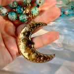 A close-up of a hand holding a gold crescent moon pendant with floral engravings, next to a beaded necklace with turquoise and gold glass beads.