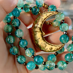 A close-up of a hand holding a beaded necklace with turquoise-blue crackled glass beads and a large gold crescent moon pendant with floral engravings.