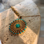 A gold sunburst pendant necklace with turquoise accents, displayed on a textured beige stone surface.