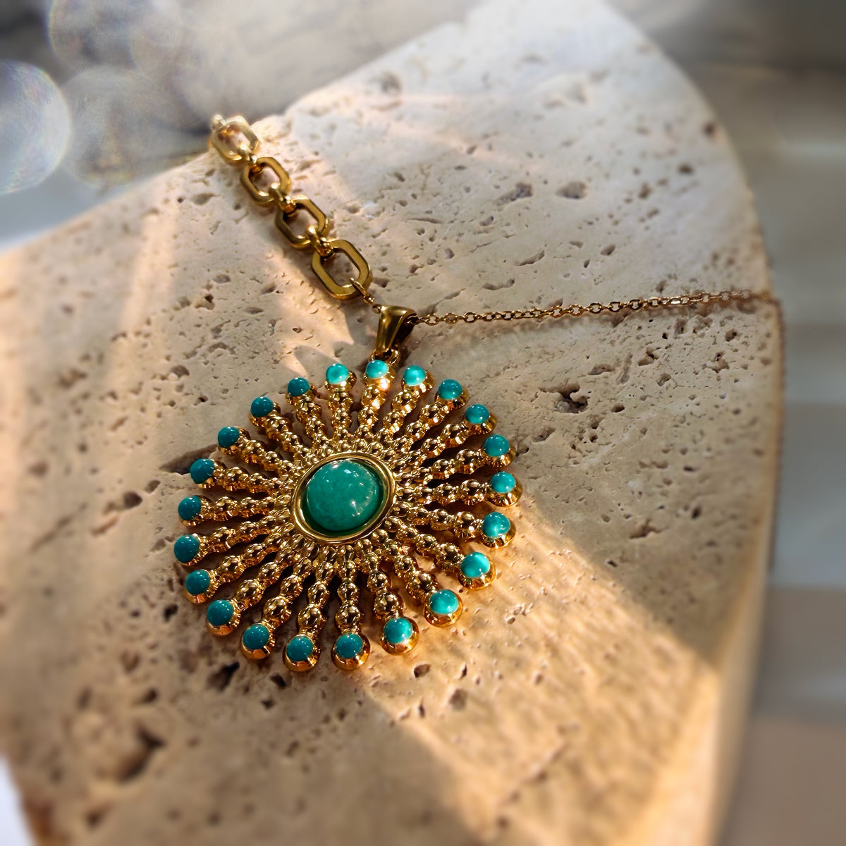 A gold sunburst pendant necklace with turquoise accents, displayed on a textured beige stone surface.