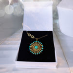 A gold pendant necklace with a turquoise center and surrounding turquoise stones, displayed on a black velvet insert inside an open white jewelry box.
