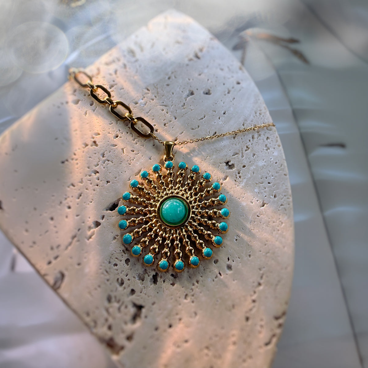 An ornate gold pendant necklace with turquoise blue accents and a central turquoise stone, displayed on a textured stone slab.