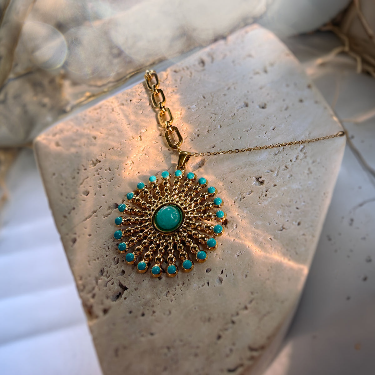 A gold-toned circular pendant necklace with a central turquoise stone and surrounding small turquoise beads, displayed on a textured stone slab.