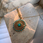 A gold-toned circular pendant necklace with a central turquoise stone and surrounding small turquoise beads, displayed on a textured stone slab.