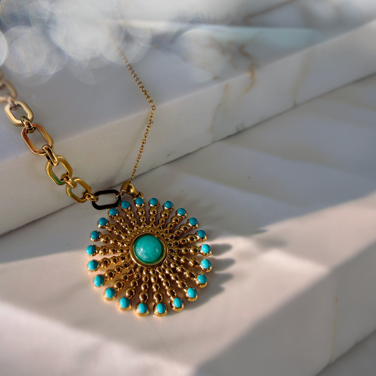 A gold-tone pendant necklace with a sunburst design, featuring turquoise-colored stones around the border and a central turquoise cabochon, resting on a white marble surface.
