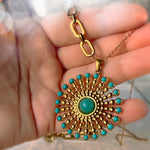 A gold-toned sunburst pendant necklace with turquoise blue accents and a central green stone, held in a person's hand.