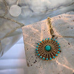 A gold-toned circular pendant necklace with a turquoise center stone and a halo of small turquoise stones, displayed on a textured stone slab with soft fabric in the background.