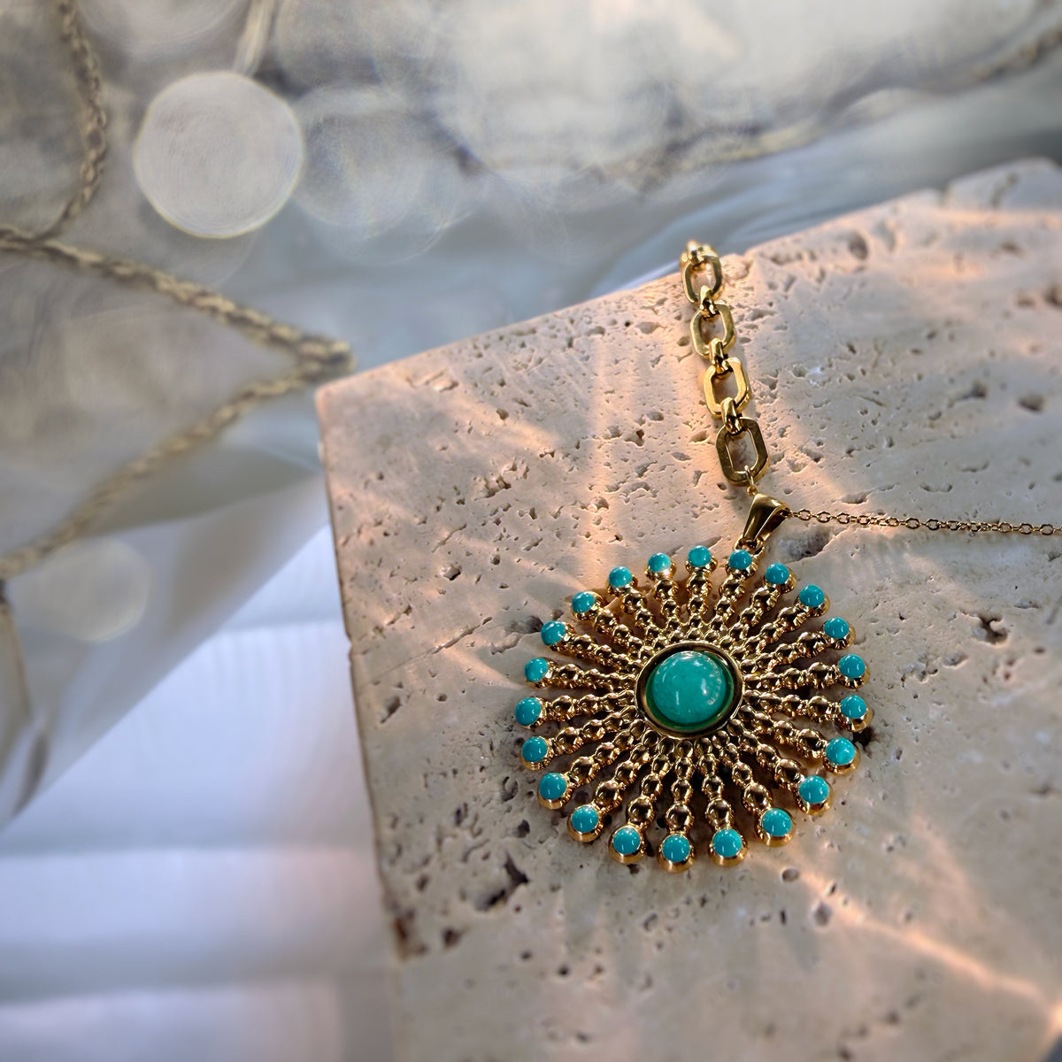 A gold-toned circular pendant necklace with a turquoise center stone and a halo of small turquoise stones, displayed on a textured stone slab with soft fabric in the background.