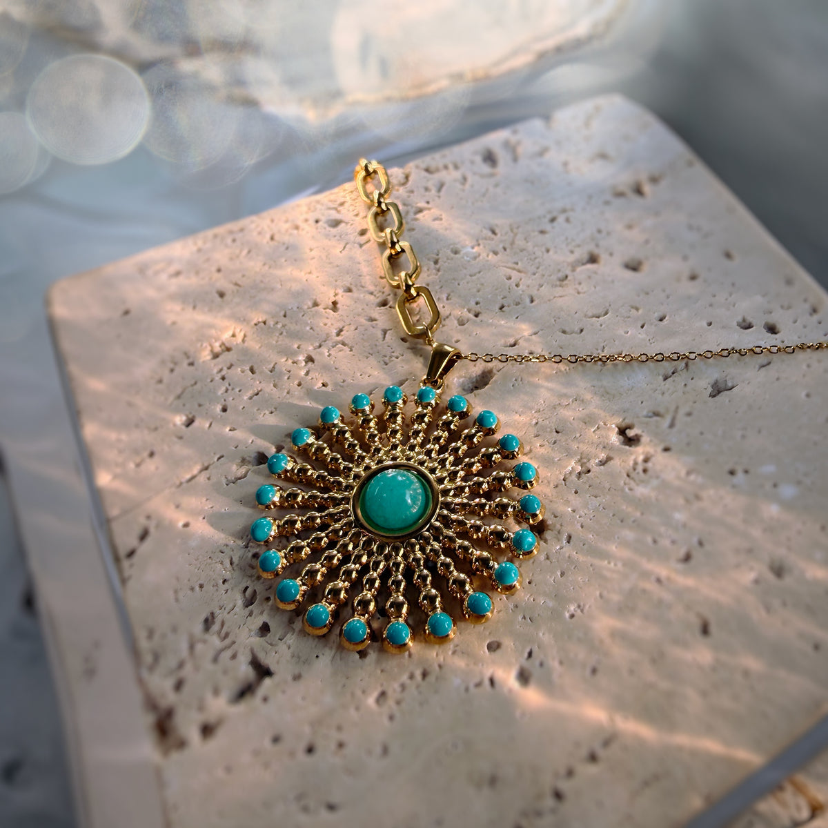 A gold-toned circular pendant necklace with turquoise blue stones, displayed on a textured stone slab with warm sunlight and bokeh in the background.