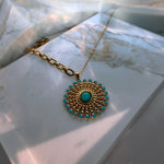 A gold pendant necklace with a sunburst design, featuring turquoise-colored stones, displayed on a white marble block.