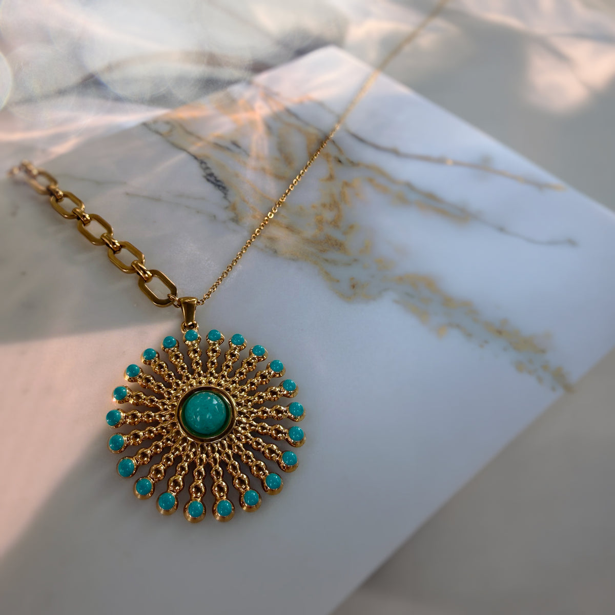 A gold-tone sunburst pendant necklace with turquoise-colored stones, displayed on a white marble card with warm sunlight and shadows.