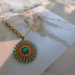 A gold-tone sunburst pendant necklace with turquoise-colored stones, displayed on a white marble card with warm sunlight and shadows.