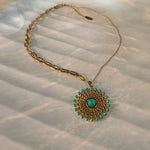 A gold chain necklace with a circular pendant featuring a central turquoise stone and surrounding small turquoise accents, displayed on a textured white fabric with soft shadows.