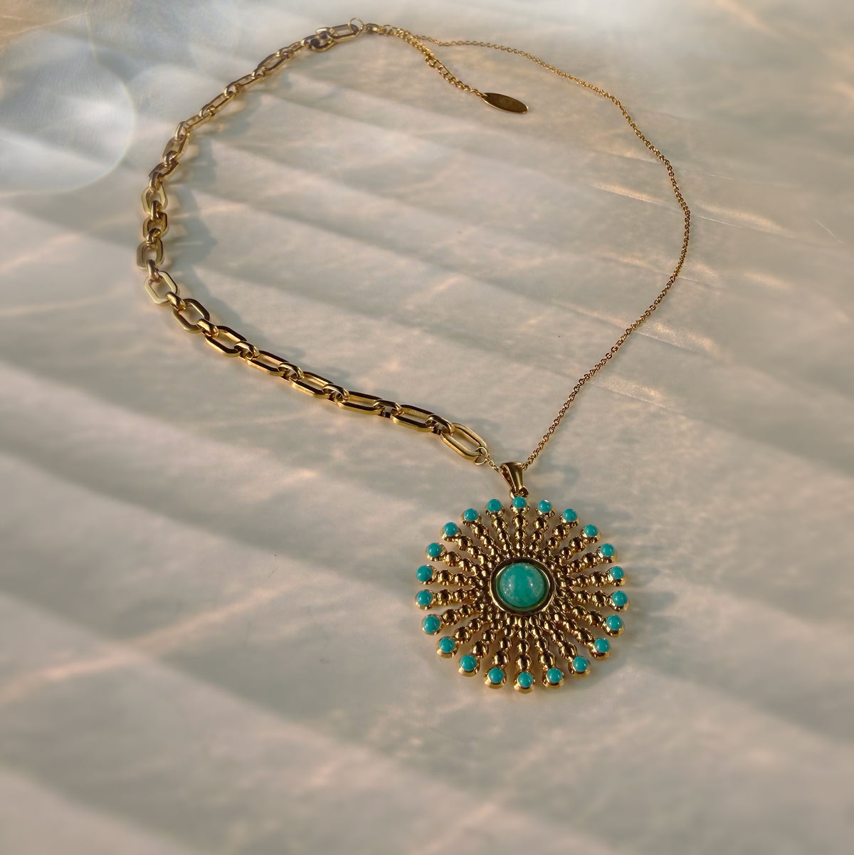 A gold chain necklace with a circular pendant featuring a central turquoise stone and surrounding small turquoise accents, displayed on a textured white fabric with soft shadows.