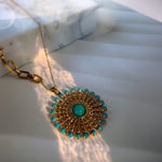 A gold-tone circular pendant necklace with a central turquoise cabochon and a border of small turquoise stones, displayed on a white marble slab with warm sunlight shadows.