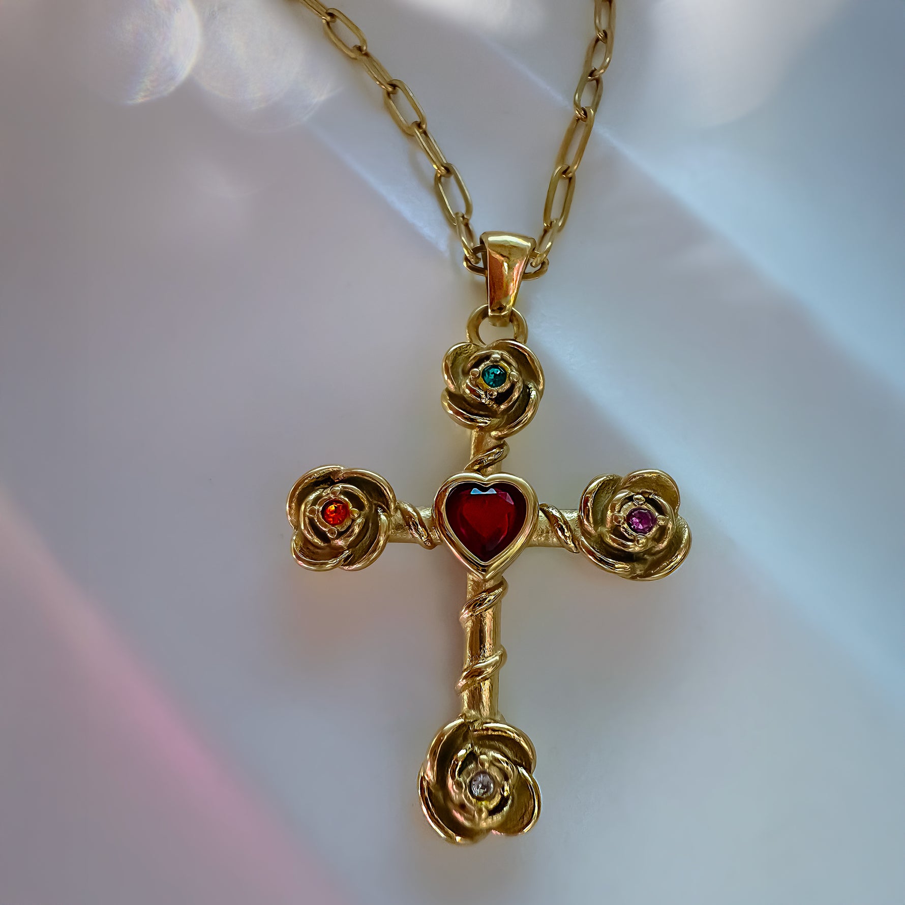 Gold cross pendant with heart and gemstones on a chain against a blurred background