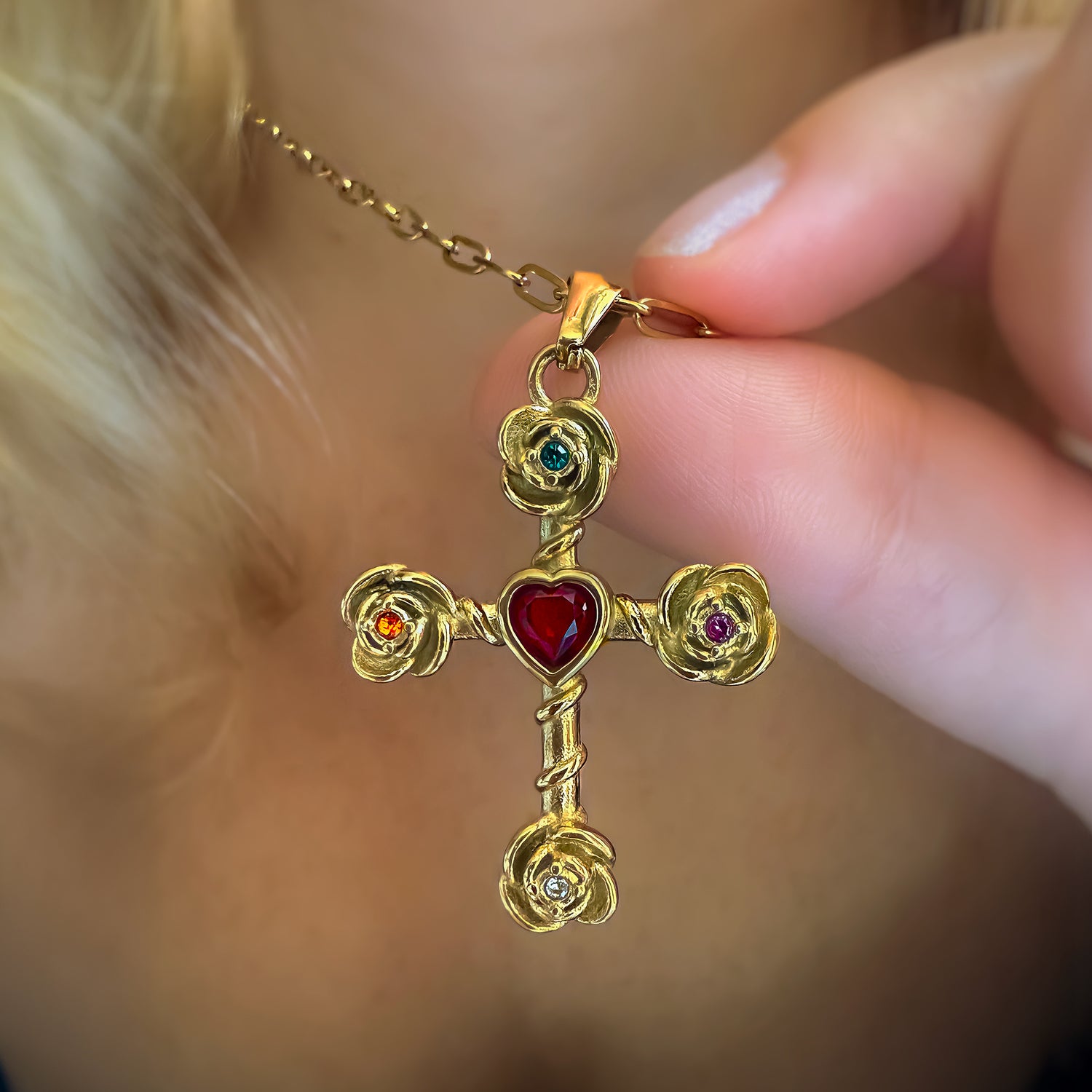 Gold cross pendant with red heart and gemstones held by a hand